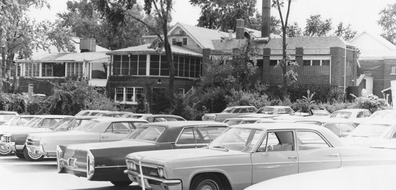 Classic cars fill a parking lot in front of a historic brick building, showcasing mid-20th century architecture and vintage vehicle culture.