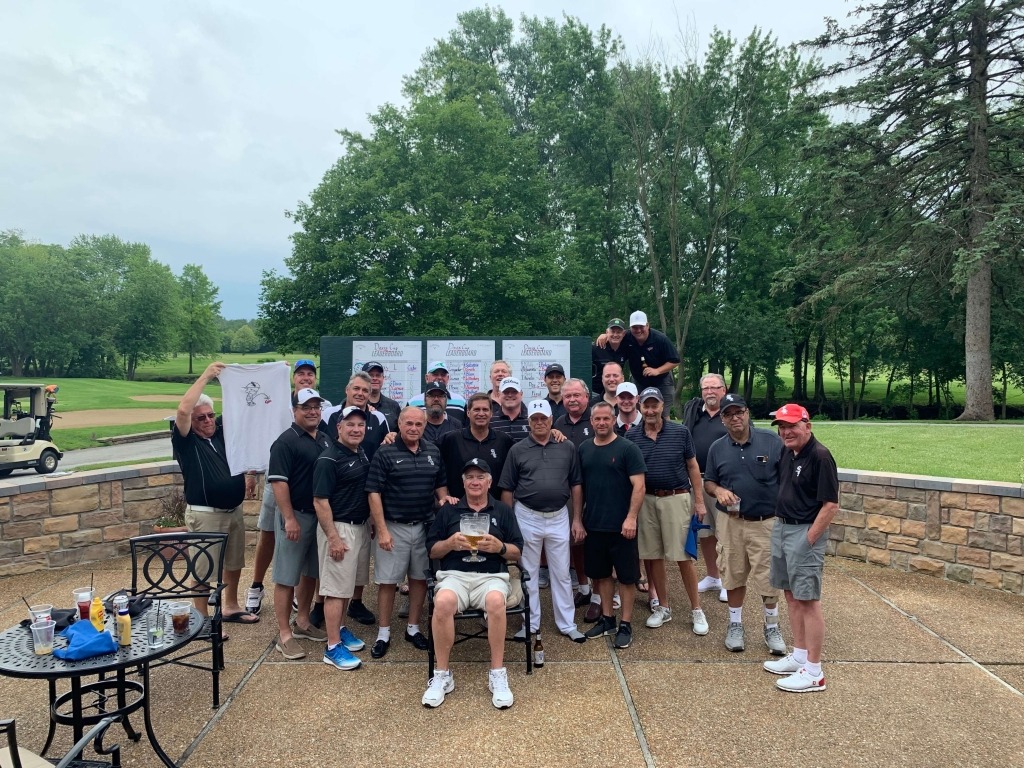 Group of golfers posed outside at a tournament, celebrating with trophies and a shirt. Lush green golf course visible in the background.