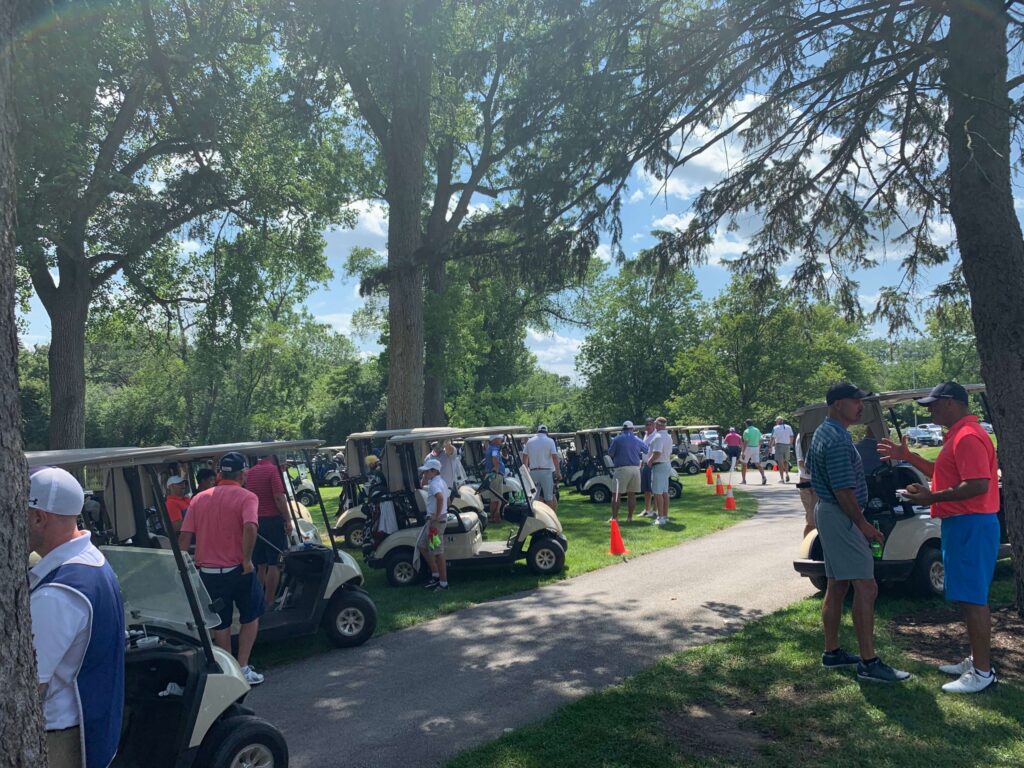 Golfers gather in the parking area, preparing for play amid numerous golf carts. A sunny day enhances the lively atmosphere of the tournament.