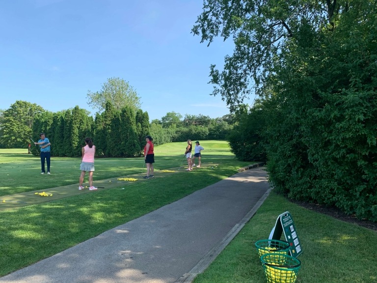 Golfers practice their swings at an outdoor driving range, surrounded by lush greenery under a clear blue sky.