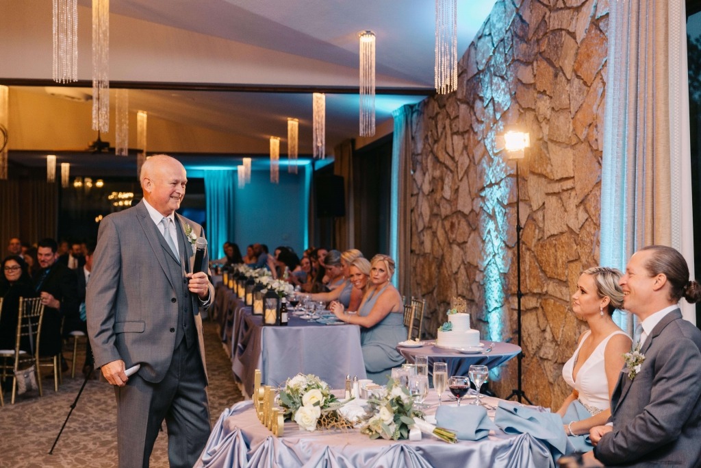 A man in a suit speaks at a wedding reception, while the bride and groom listen attentively. Guests enjoy the celebration in a decorated venue.