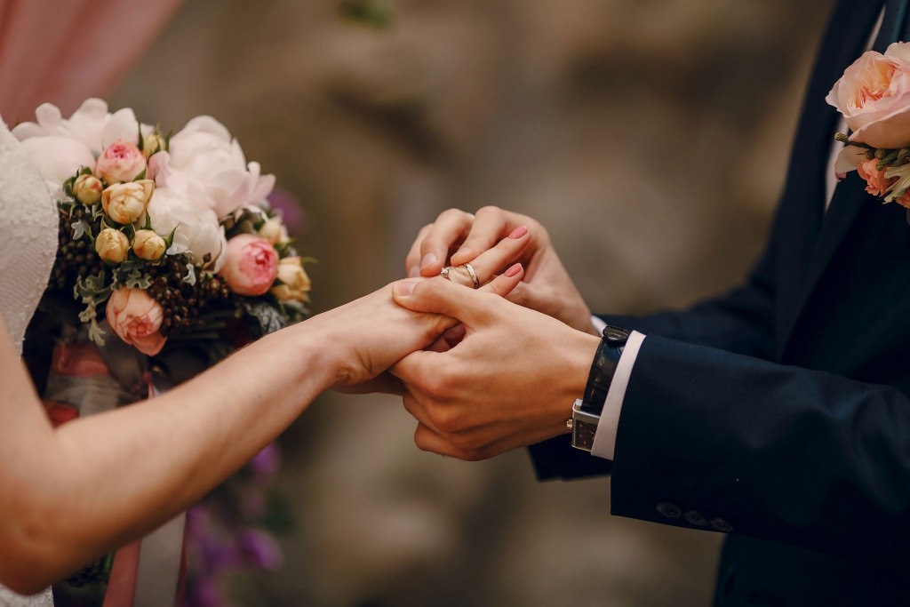 A bride and groom exchange wedding rings, symbolizing their commitment, with a bouquet of pink and white flowers in the bride’s hand.