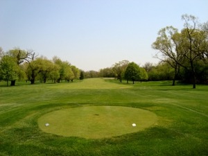 A well-maintained golf course with a clear view of the fairway and greens, surrounded by lush trees and a bright blue sky.