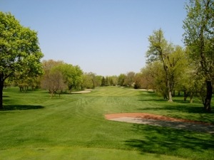 Lush golf course fairway lined with trees under a clear blue sky, featuring a sand trap on the right, emphasizing a serene outdoor setting.