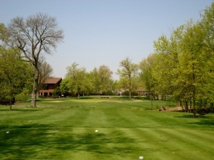 Lush green golf course fairway lined with trees, leading to a club building in the background under a clear blue sky.